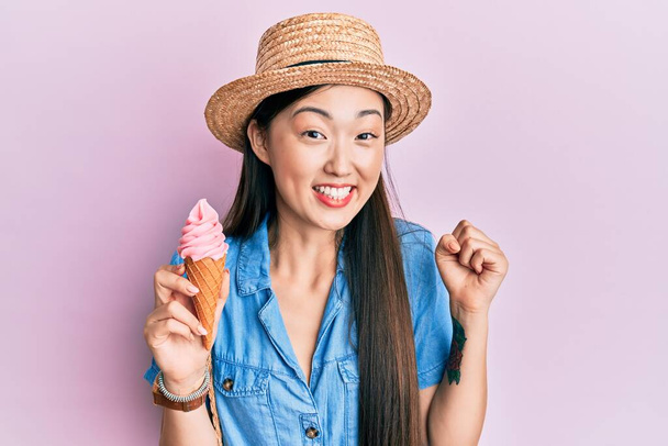 Young chinese woman wearing summer hat holding ice cream screaming proud, celebrating victory and success very excited with raised arm  - Фото, зображення