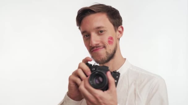 Caucasian bearded brunette man wear shirt smile, hold retro photo camera, take a shot isolated on white wall background close up. Photographer guy kissed by girlfriend, red lipstick kiss marks on face - Footage, Video