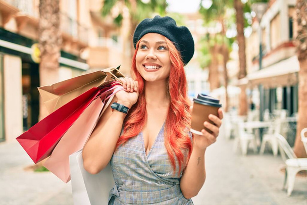 Młoda ruda dziewczyna nosi francuski styl trzymając torby na zakupy i picia zabrać kawę w mieście. - Zdjęcie, obraz
