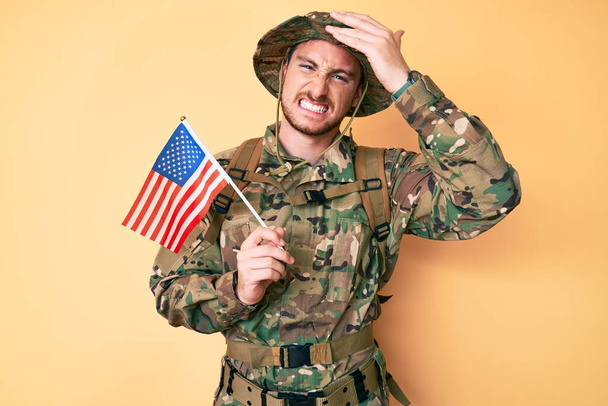 Junger kaukasischer Mann in Tarnuniform mit einer US-Fahne auf dem Kopf, überrascht und wütend  - Foto, Bild