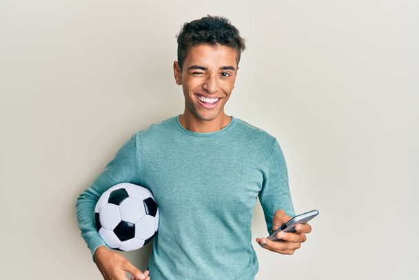 Νεαρός όμορφος Αφροαμερικάνος που κρατάει μπάλα ποδοσφαίρου κοιτάζοντας το smartphone να κλείνει το μάτι κοιτάζοντας την κάμερα με σέξι έκφραση, χαρούμενο και χαρούμενο πρόσωπο.  - Φωτογραφία, εικόνα