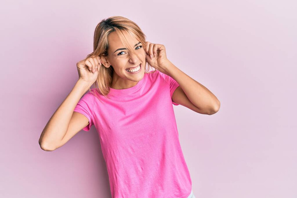 Bella donna bionda che indossa casual t-shirt rosa sorridente tirando le orecchie con le dita, gesto divertente. problema di audizione  - Foto, immagini