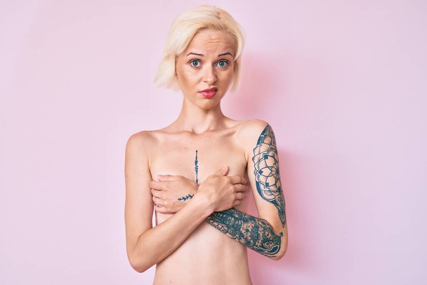 Jonge blonde vrouw met tatoeage staan shirtloze clueless en verwarde uitdrukking. twijfelconcept.  - Foto, afbeelding