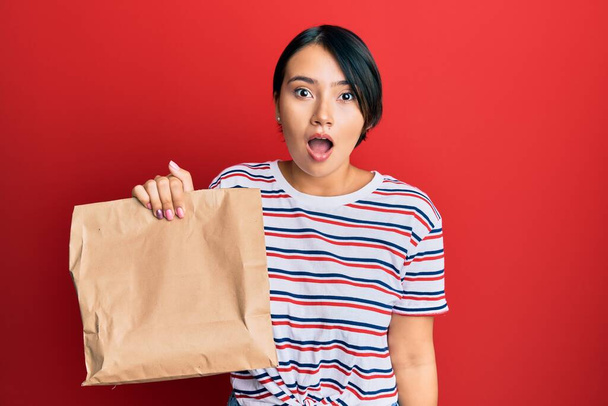 Beautiful young woman with short hair holding take away paper bag scared and amazed with open mouth for surprise, disbelief face  - Foto, Imagem