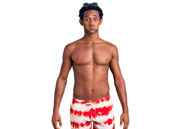 African handsome man wearing swimsuit and sunglasses puffing cheeks with funny face. mouth inflated with air, crazy expression.  - Фото, изображение