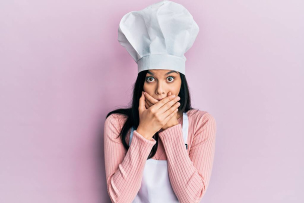 Mulher hispânica jovem vestindo uniforme padeiro e chapéu cozinheiro chocado cobrindo a boca com as mãos por engano. conceito secreto.  - Foto, Imagem
