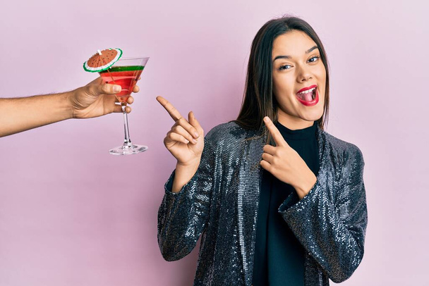 Young girl wearing sexy party jacket and hispanic arm offering cocktail smiling and looking at the camera pointing with two hands and fingers to the side.  - Φωτογραφία, εικόνα
