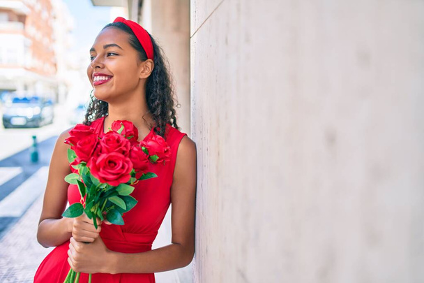 Mladý africký americký dívka usměvavý šťastný držení kytice růží opírající se o zeď ve městě. - Fotografie, Obrázek