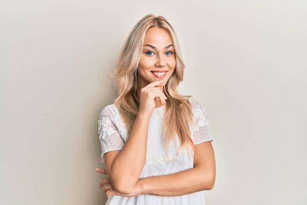 Όμορφη καυκάσια ξανθιά κοπέλα φορώντας λευκό πουκάμισο χαμογελώντας αναζητούν αυτοπεποίθηση στην κάμερα με σταυρωμένα χέρια και το χέρι στο πηγούνι. θετική σκέψη.  - Φωτογραφία, εικόνα