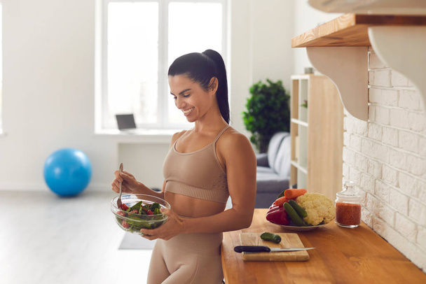 Femme de fitness mince en vêtements de sport debout à la maison dans la cuisine avec un bol de salade fraîche. - Photo, image
