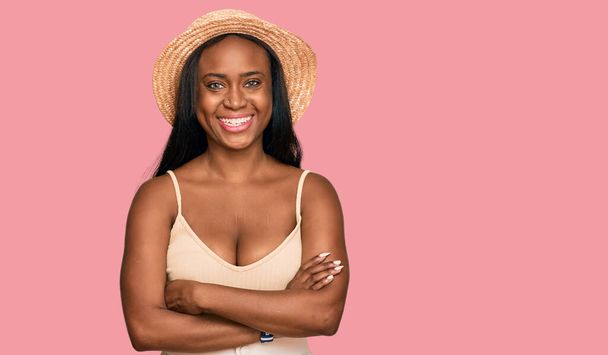 Jonge zwarte vrouw met zomerhoed vrolijk gezicht glimlachend met gekruiste armen kijkend naar de camera. positieve persoon.  - Foto, afbeelding