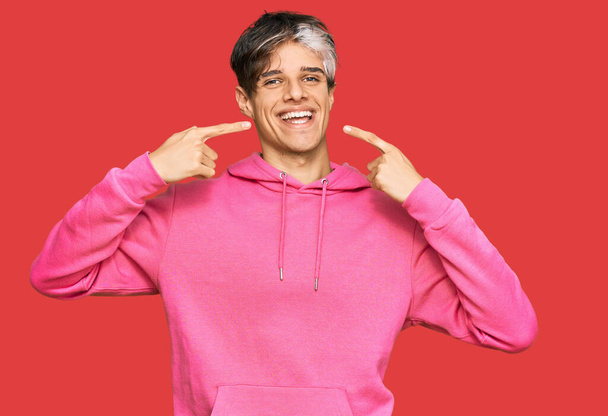 Młody Latynos w luźnej różowej bluzie, uśmiechnięty, radosny, pokazywany palcami, zębami i ustami. Koncepcja zdrowia zębów.  - Zdjęcie, obraz