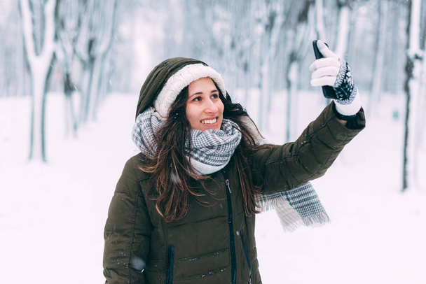 Gülümseyen genç kadın karla kaplı bir ormanda selfie çekiyor. - Fotoğraf, Görsel