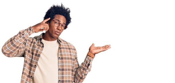 Bello uomo afro-americano con i capelli afro indossa abiti casual e occhiali confusi e infastiditi con palmo aperto mostrando spazio copia e puntando il dito sulla fronte. Pensaci..  - Foto, immagini
