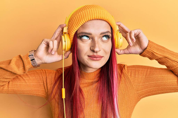 ヘッドフォンを使って音楽を聴いている若い白人女性は、横を見て考えずに笑っている。.  - 写真・画像