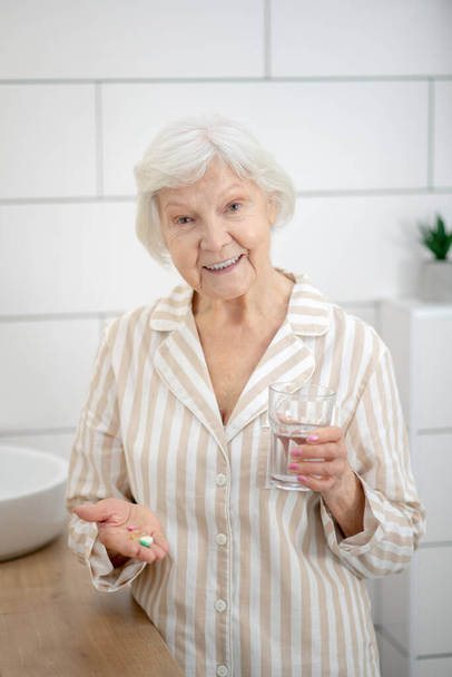 Woman in pajama holding a pills in her hands and a glass of water - Fotoğraf, Görsel