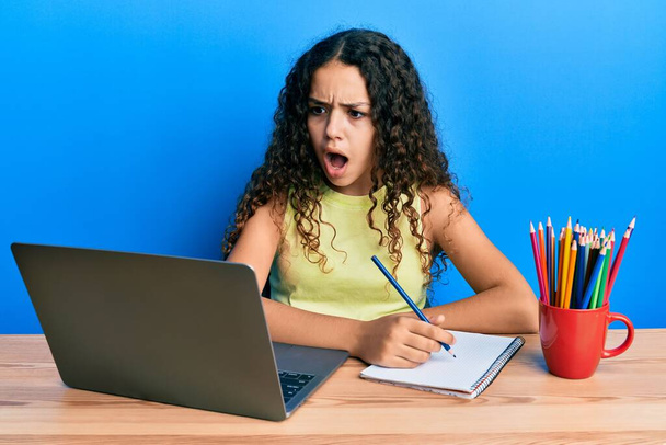 Teenager hispanic girl sitting on the table studying for school angry and mad screaming frustrated and furious, shouting with anger. rage and aggressive concept.  - Fotó, kép