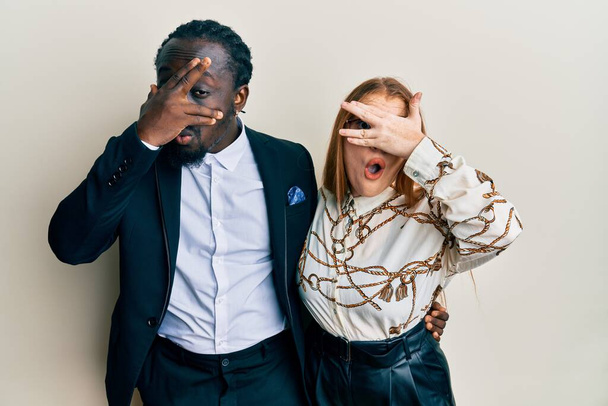 Young interracial couple wearing business and elegant clothes peeking in shock covering face and eyes with hand, looking through fingers with embarrassed expression.  - Photo, Image