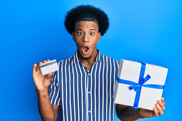 Африканский американец с афроволосами держит подарок и кредитную карту боится и шокирован удивлением и удивлением выражение лица, страх и возбужденное лицо.  - Фото, изображение