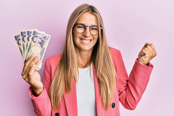 Young blonde woman wearing business style holding japanese yen banknotes screaming proud, celebrating victory and success very excited with raised arm  - Foto, afbeelding