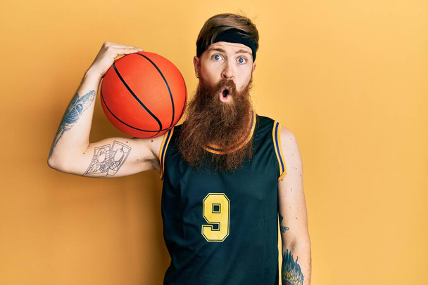 Рыжий мужчина с длинной бородой в баскетбольной форме держит мяч испуганный и удивленный с открытым ртом для удивления, неверие лицо  - Фото, изображение