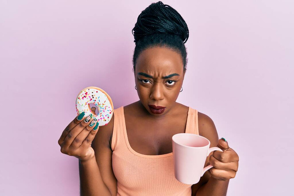 Jeune femme afro-américaine mangeant des beignets et buvant du café expression désorientée et confuse. concept de doute.  - Photo, image