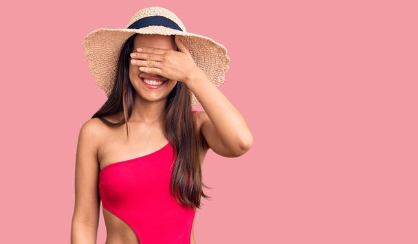 Junge schöne lateinische Mädchen in Badebekleidung und Sommermütze lächeln und lachen mit der Hand auf dem Gesicht bedecken Augen für die Überraschung. Blindes Konzept.  - Foto, Bild