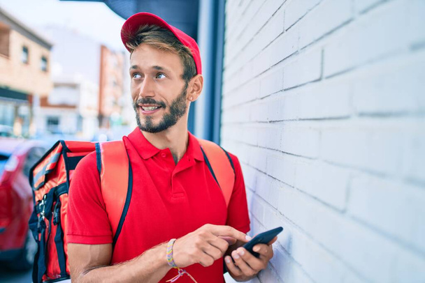 Kaukaski dostawca w czerwonym mundurze i plecak dostawy uśmiecha się szczęśliwy na zewnątrz za pomocą smartfona - Zdjęcie, obraz