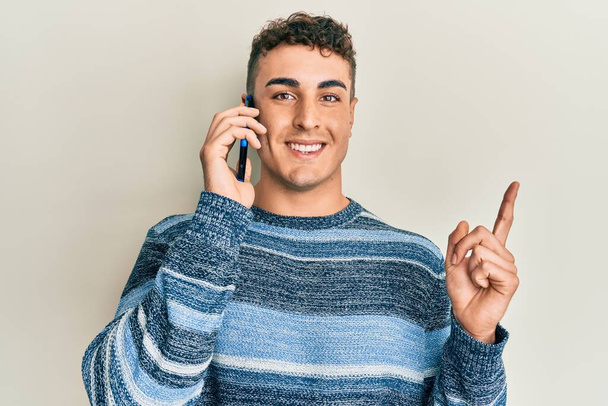 Joven hispano conversando hablando en el smartphone sonriendo feliz señalando con la mano y el dedo hacia un lado  - Foto, imagen