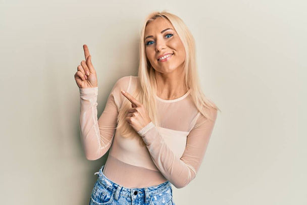 Mladá blondýna žena na sobě ležérní oblečení s úsměvem a při pohledu na kameru ukazuje dvěma rukama a prsty na stranu.  - Fotografie, Obrázek