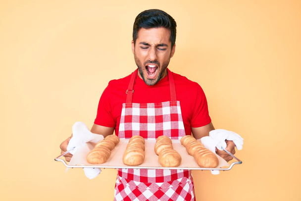 Young hispanic man wearing baker apron holding homemade bread angry and mad screaming frustrated and furious, shouting with anger. rage and aggressive concept.  - Foto, afbeelding