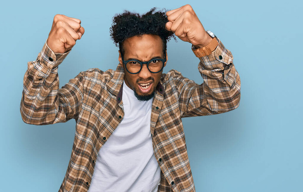 Young african american man with beard wearing casual clothes and glasses angry and mad raising fists frustrated and furious while shouting with anger. rage and aggressive concept.  - 写真・画像