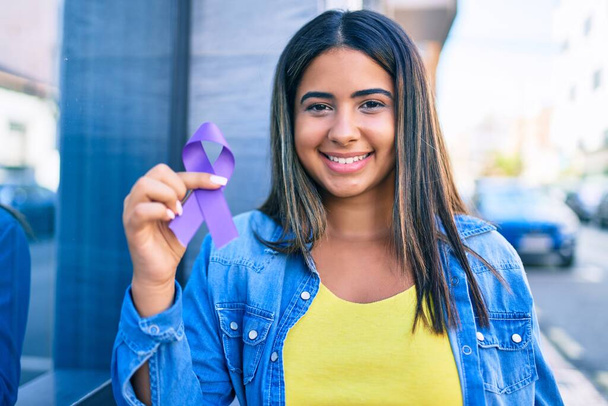 Молодая латинская женщина улыбается счастливо держа фиолетовую ленту в городе. - Фото, изображение