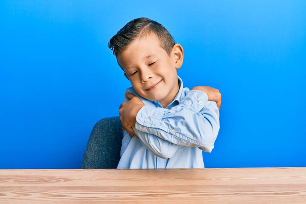 Adorable enfant caucasien portant des vêtements décontractés assis sur la table s'embrassant heureux et positif, souriant confiant. amour de soi et soins de soi  - Photo, image