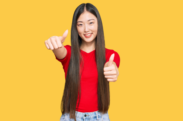 Mladá Číňanka, oblečená do neformálního oblečení a schvalující pozitivní gesto rukou, s palcem nahoru, usmívajícím se a šťastným za úspěch. gesto vítěze.  - Fotografie, Obrázek