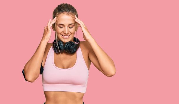 Young blonde woman wearing gym clothes and using headphones with hand on head for pain in head because stress. suffering migraine.  - Photo, image
