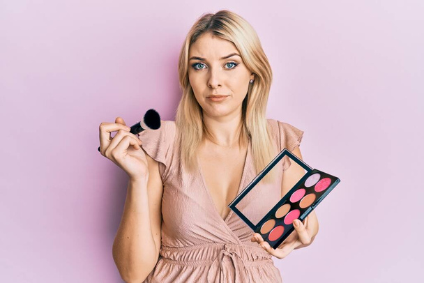 Young caucasian woman holding makeup brush and blush palette skeptic and nervous, frowning upset because of problem. negative person.  - Photo, Image