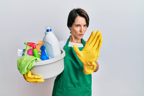 Young brunette woman with short hair wearing apron holding cleaning products doing stop gesture with hands palms, angry and frustration expression  - Φωτογραφία, εικόνα