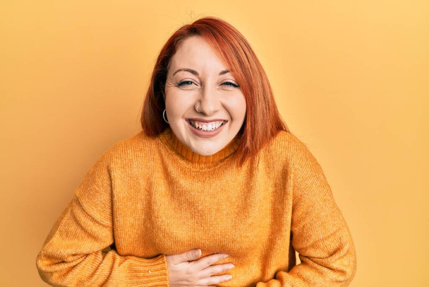 Hermosa mujer pelirroja vistiendo suéter de invierno casual sobre fondo amarillo sonriendo y riendo en voz alta porque divertida broma loca con las manos en el cuerpo.  - Foto, Imagen