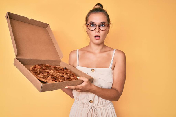 Mulher branca bonita segurando pizza de pepperoni saborosa no rosto de choque, olhando cético e sarcástico, surpreso com a boca aberta  - Foto, Imagem