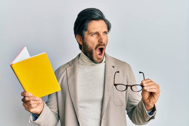 Young hispanic man reading a book and holding glasses angry and mad screaming frustrated and furious, shouting with anger. rage and aggressive concept.  - Photo, Image
