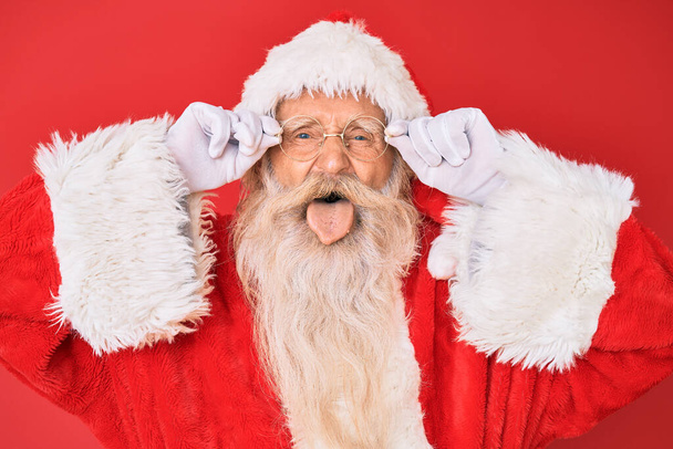 Old senior man with grey hair and long beard wearing santa claus costume holding glasses sticking tongue out happy with funny expression.  - Photo, Image