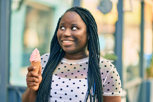 Joven mujer afroamericana sonriendo feliz comiendo helado en la ciudad. - Foto, imagen