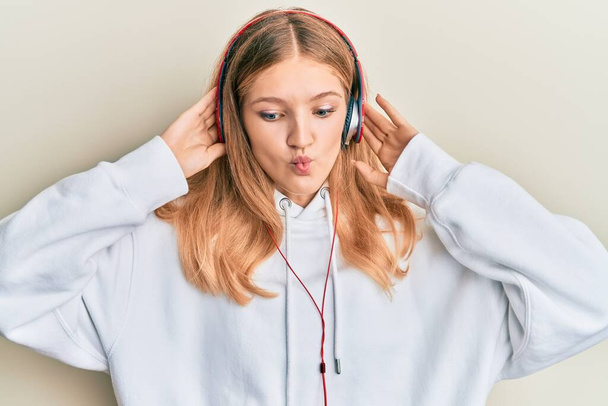 Όμορφη νεαρή καυκάσια κοπέλα ακούγοντας μουσική χρησιμοποιώντας ακουστικά κάνοντας το πρόσωπο ψαριών με το στόμα και αλληθωρίζοντας τα μάτια, τρελό και κωμικό.  - Φωτογραφία, εικόνα
