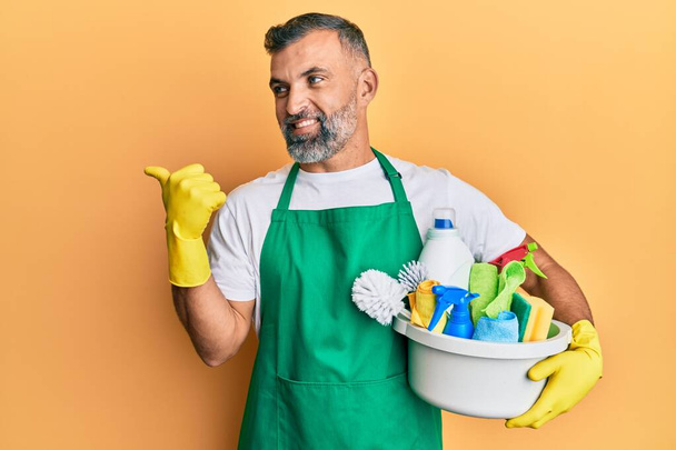 Middle age handsome man holding cleaning products pointing thumb up to the side smiling happy with open mouth  - Fotografie, Obrázek