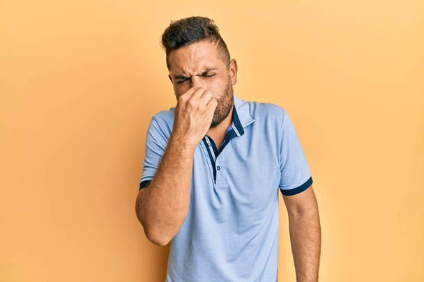 Handsome man with beard wearing casual clothes smelling something stinky and disgusting, intolerable smell, holding breath with fingers on nose. bad smell  - Photo, Image