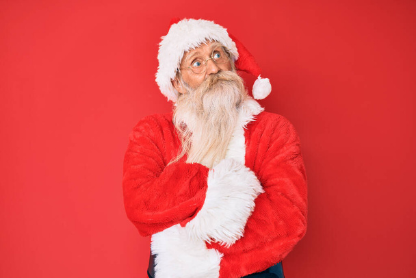 Vecchio uomo anziano con i capelli grigi e la barba lunga che indossa il costume tradizionale di Babbo Natale sorridente guardando di lato e fissando via pensando.  - Foto, immagini