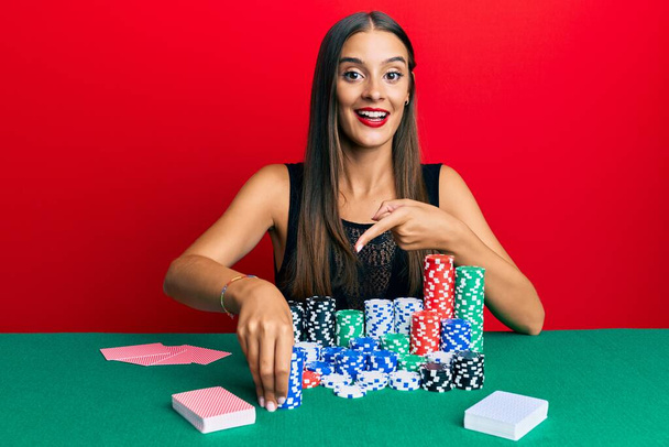 Mujer hispana joven sentada en la mesa sosteniendo fichas de casino sonriendo feliz señalando con la mano y el dedo  - Foto, Imagen