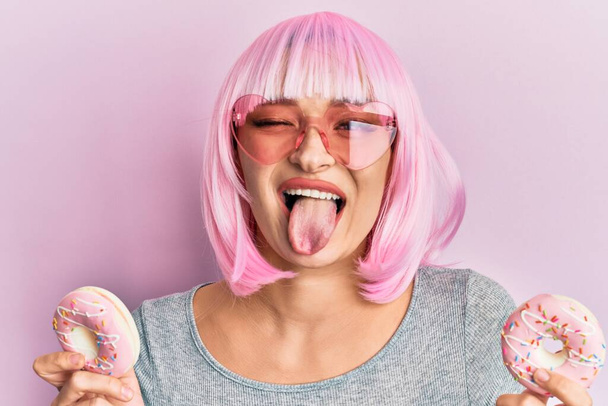 Junge kaukasische Frau mit pinkfarbener Perücke und Donuts, die die Zunge herausstrecken, mit lustigem Ausdruck glücklich.  - Foto, Bild