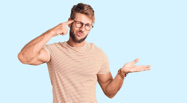 Young caucasian man wearing casual clothes and glasses confused and annoyed with open palm showing copy space and pointing finger to forehead. think about it.  - Photo, Image